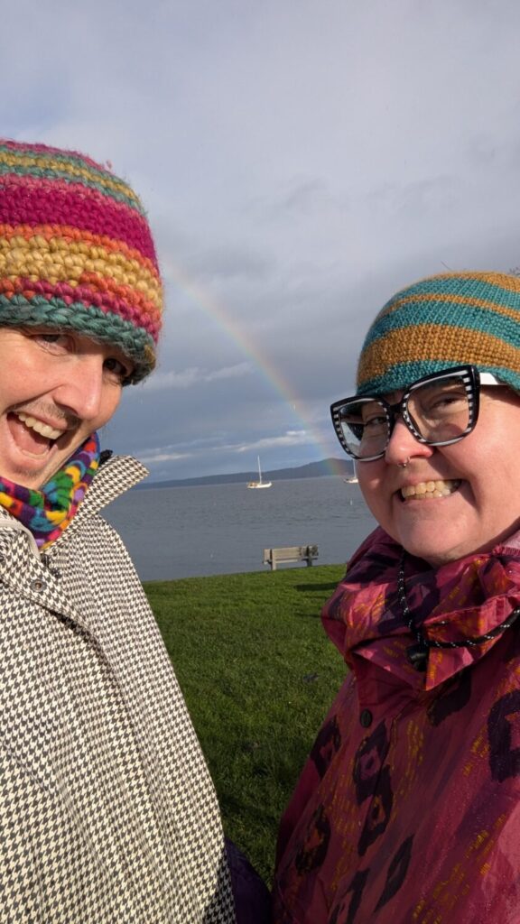 Kori & Jess in rain wear and home made toques smile exaggeratedly under a big rainbow over the beach.